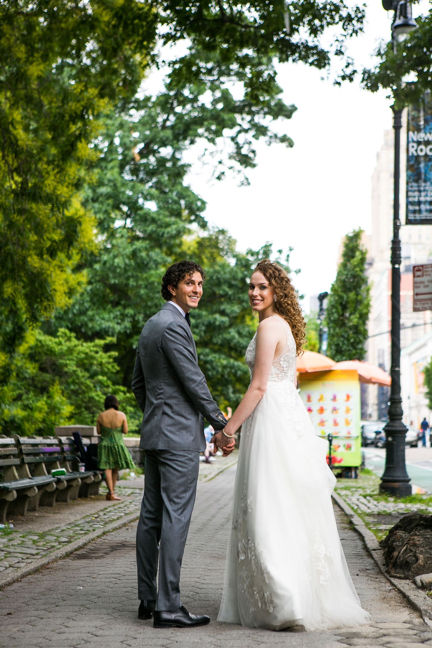 Bride and groom holding hands on a tree-lined city sidewalk, turning to smile at the camera.