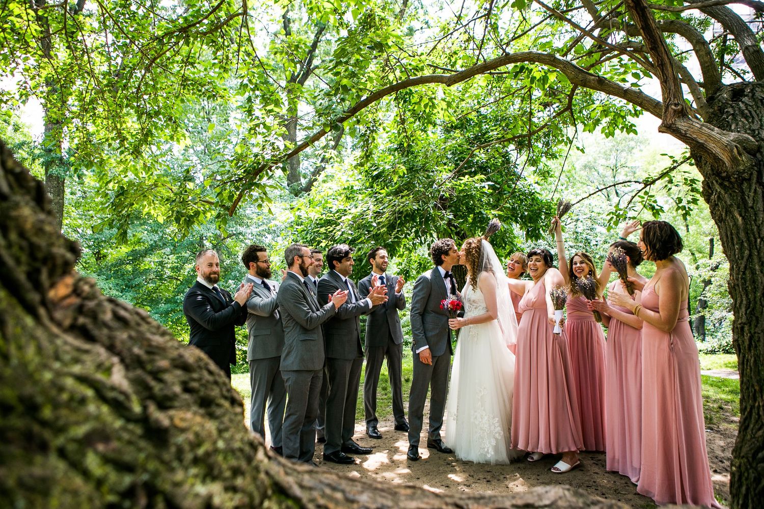 Wedding party laughing and celebrating under a curved tree canopy with pink bridesmaids and gray suits.