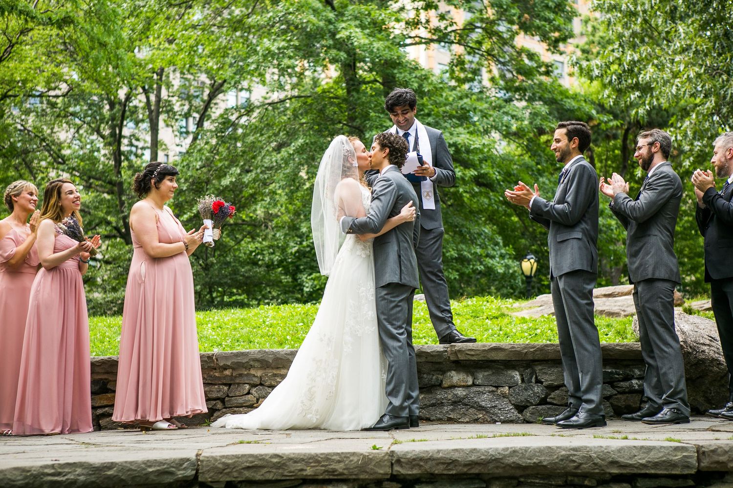 Bride and groom share first kiss at outdoor wedding ceremony with bridesmaids in pink dresses and groomsmen in gray suits.