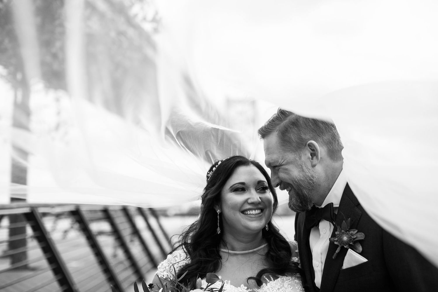 Black and white close-up of laughing bride and groom as veil swirls around them outdoors.