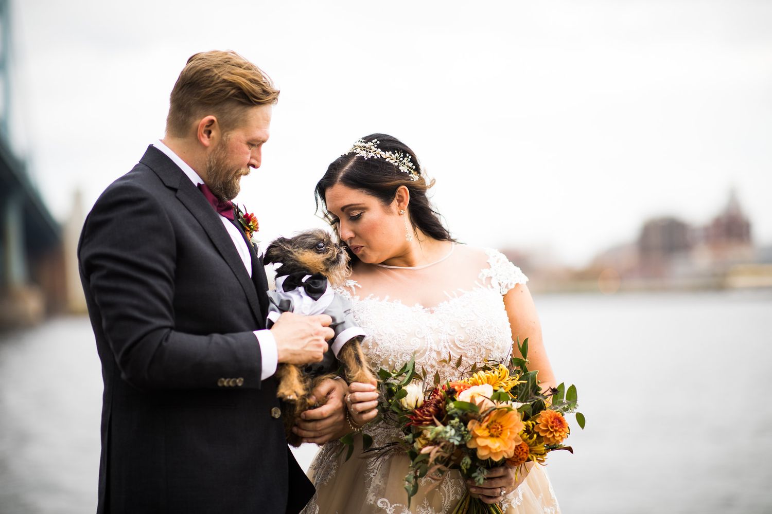 Bride and groom tenderly hold a small dressed dog by the river, bride carrying an autumn bouquet.