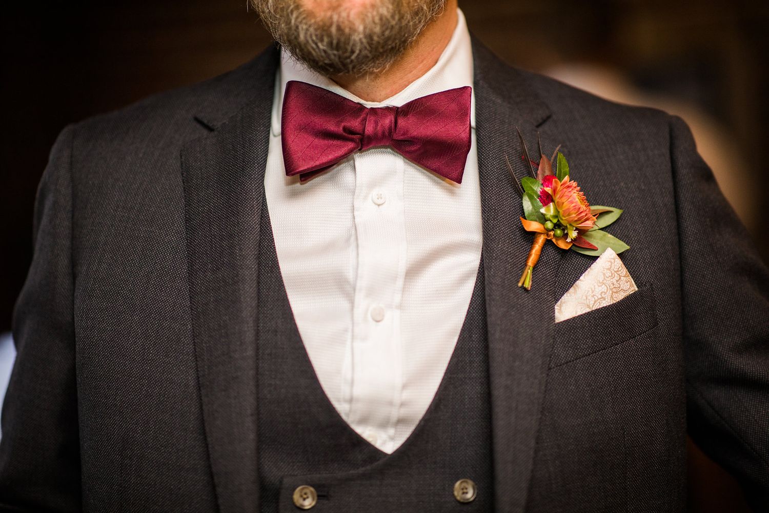 Close-up of groom's burgundy bow tie, boutonniere, and pocket square on dark suit.