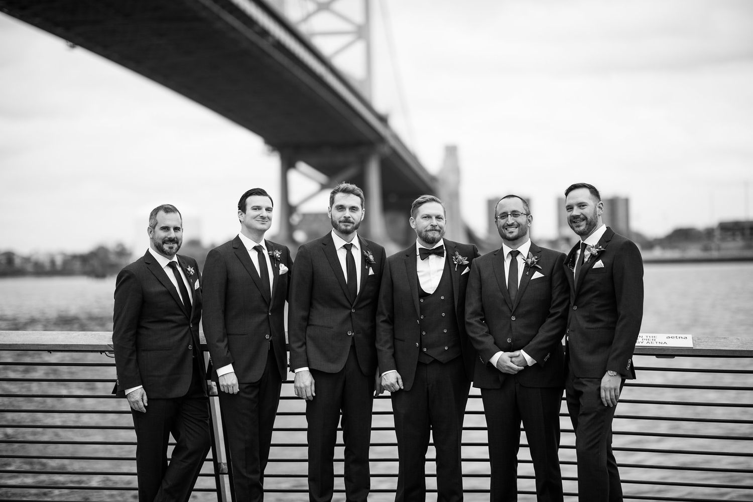 Black and white photo of a groom and five groomsmen in dark suits posing by a river with a bridge behind them.