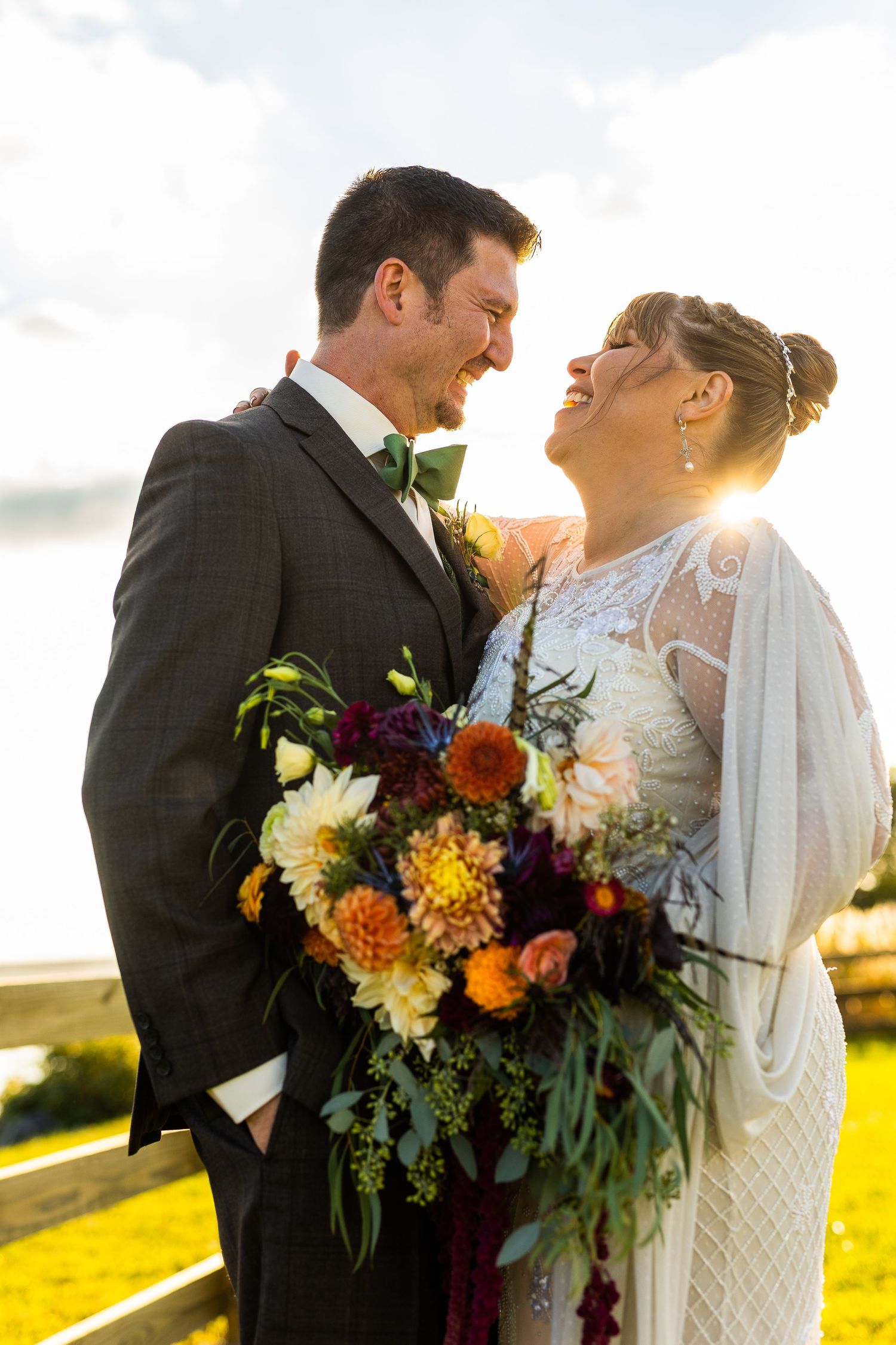 Bride and groom laughing together at golden hour, bride holding a vibrant orange and burgundy bouquet.