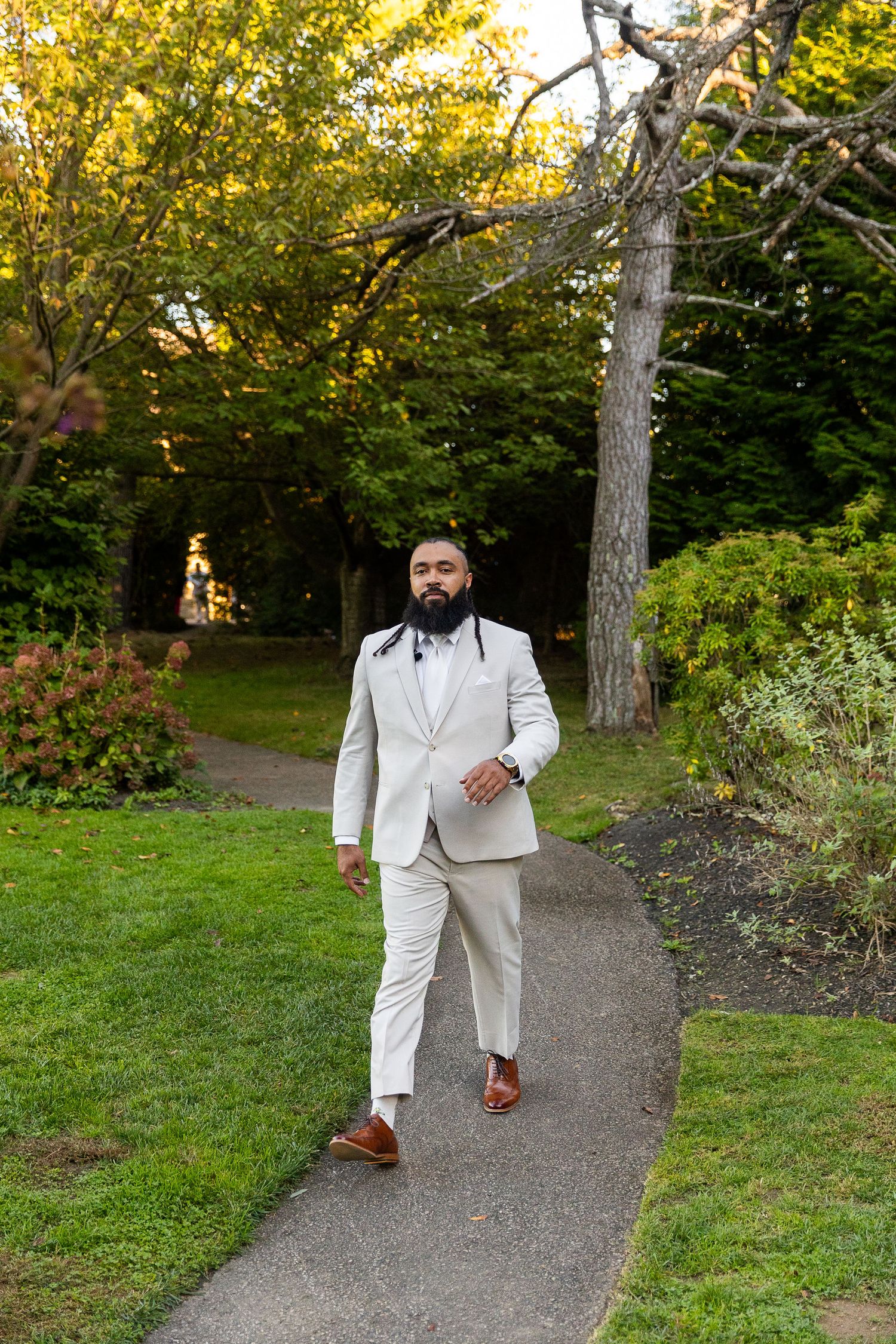 Bearded groom in beige suit walking confidently along garden path at golden hour.
