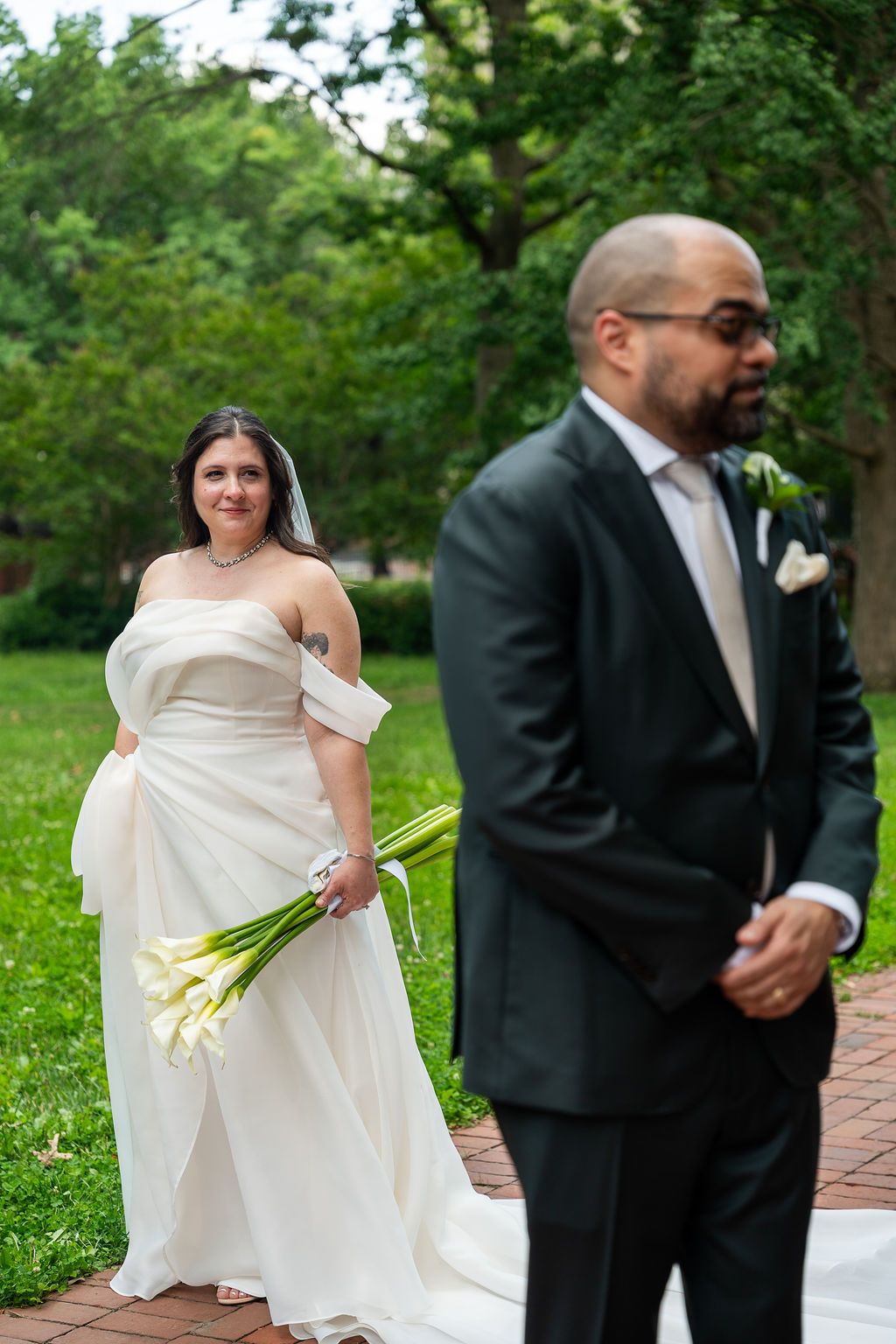 Bride in white strapless gown with calla lilies smiles as groom stands in foreground in dark suit.