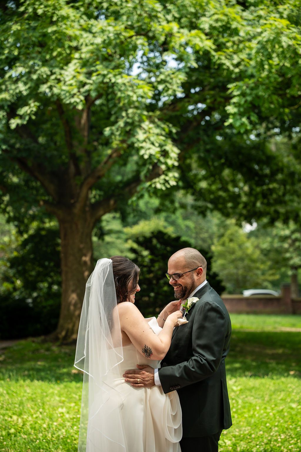 Smiling groom in dark suit holds bride close during outdoor wedding portraits in a lush green park.