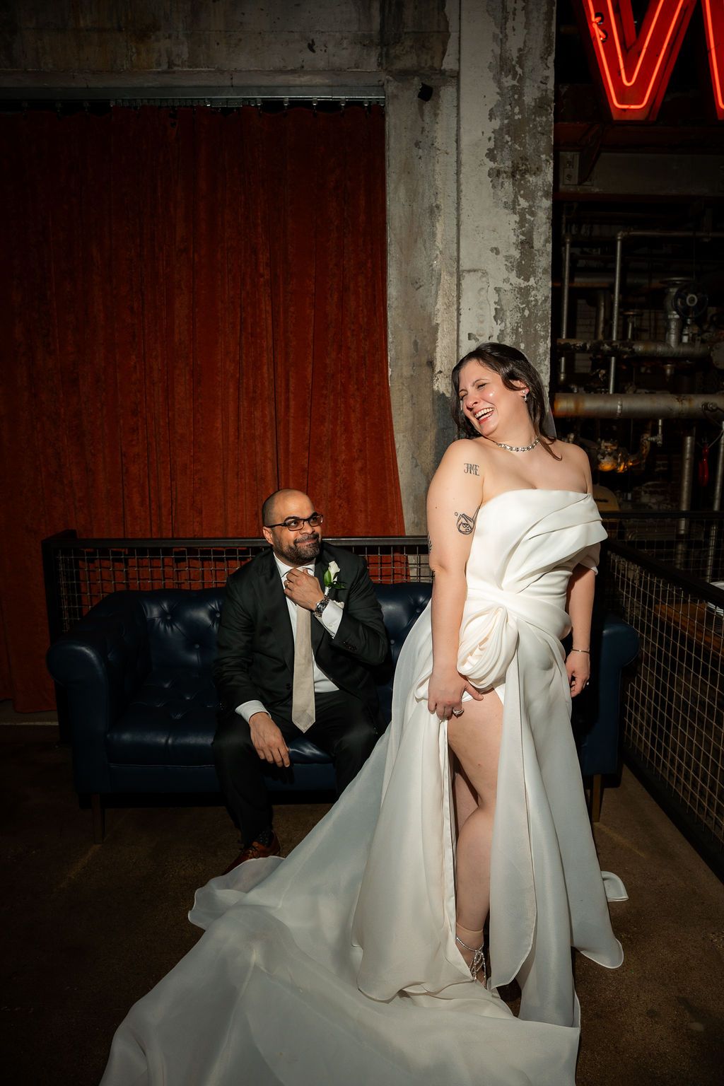 Joyful bride in off-shoulder white gown laughing while groom smiles from blue couch under neon sign.