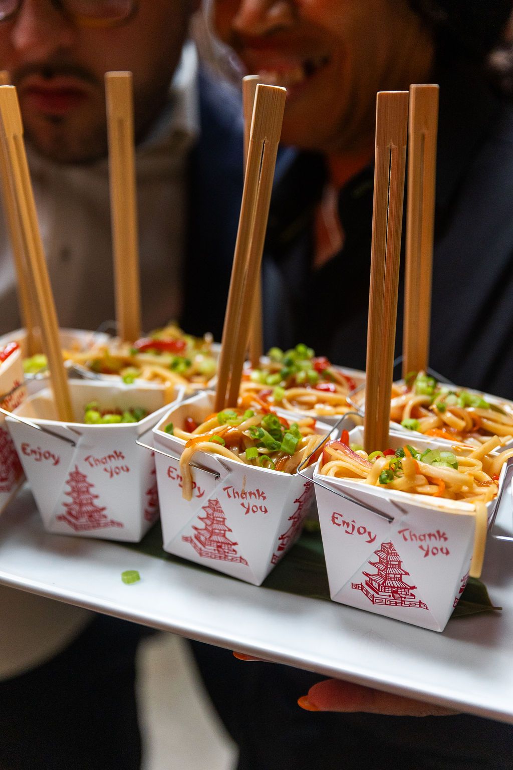 Server presenting a tray of Chinese takeout boxes filled with noodles and chopsticks at a catered party.