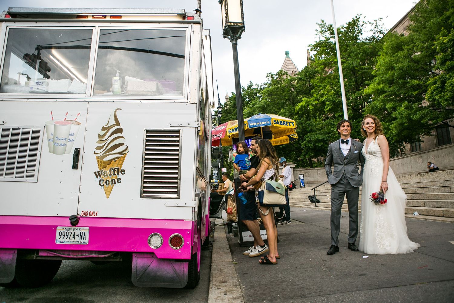 Bride and groom posing beside a pink and white Wafels and Dinges food truck on a sunny city street.