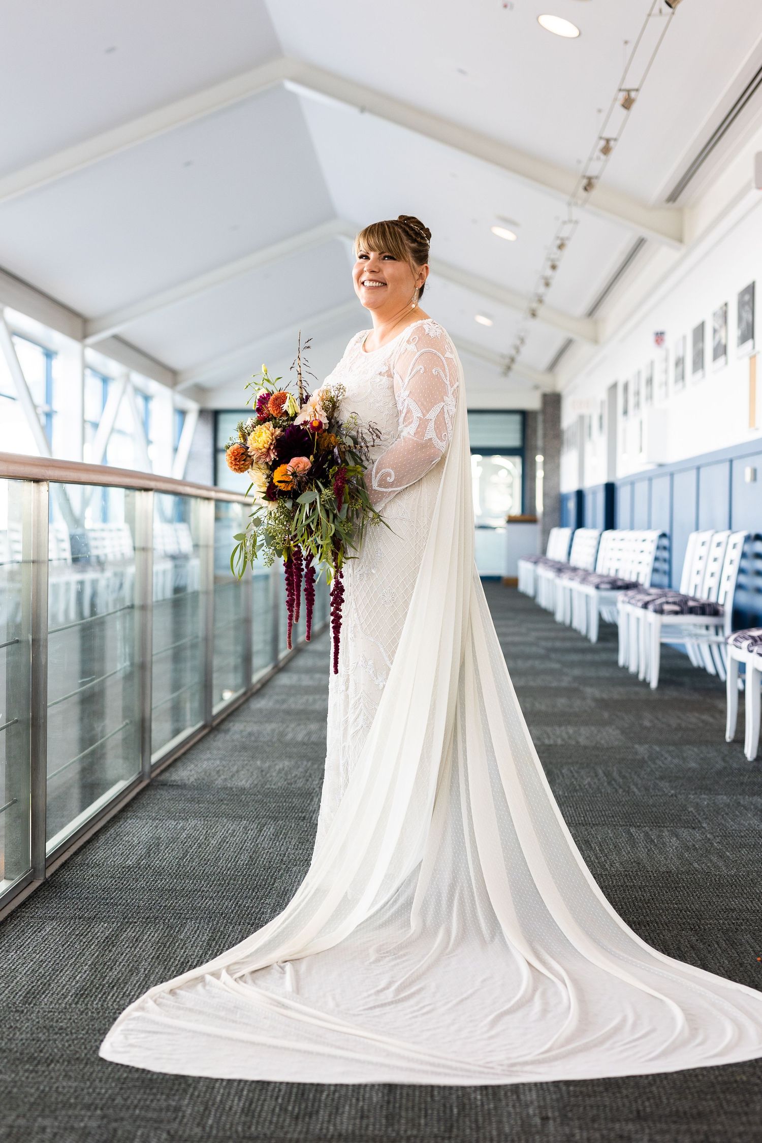 Smiling bride in long-sleeve lace wedding gown with flowing train holding vibrant fall bouquet indoors.