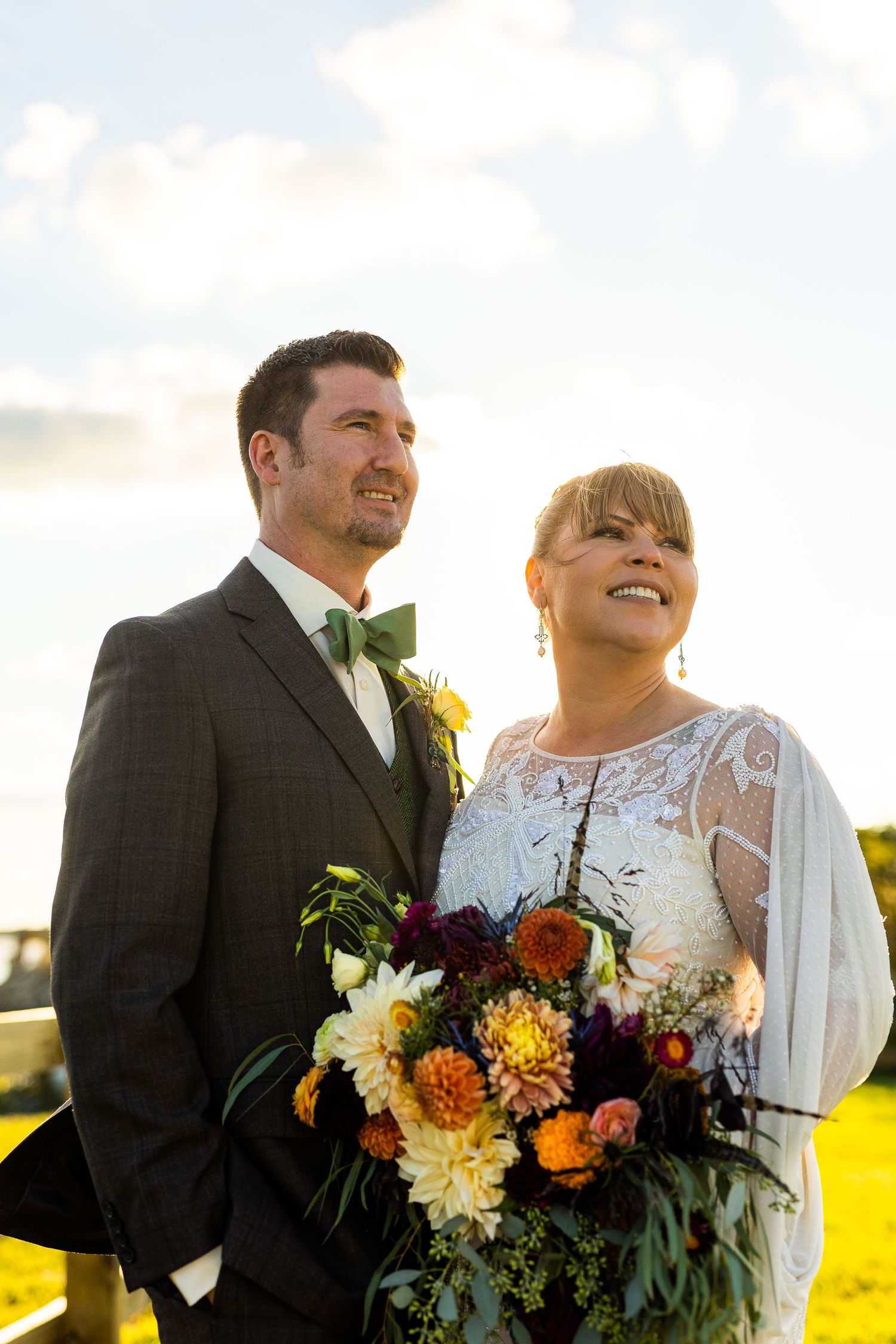 Bride and groom smiling and looking up outdoors at golden hour, bride holding a colorful floral bouquet.