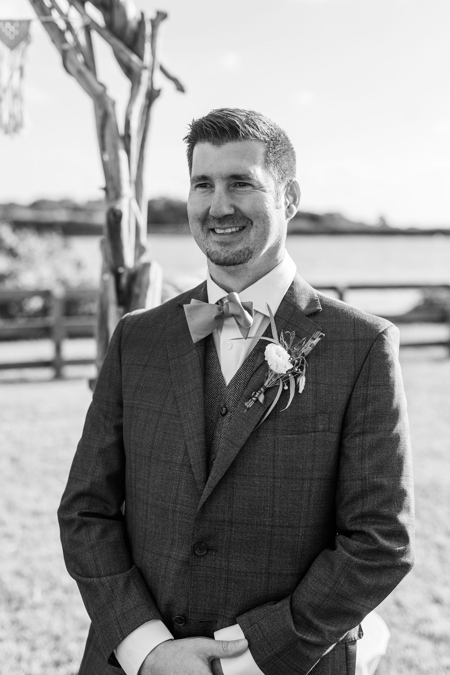 Black and white close-up portrait of a smiling groom in a plaid suit with boutonniere at an outdoor ceremony.