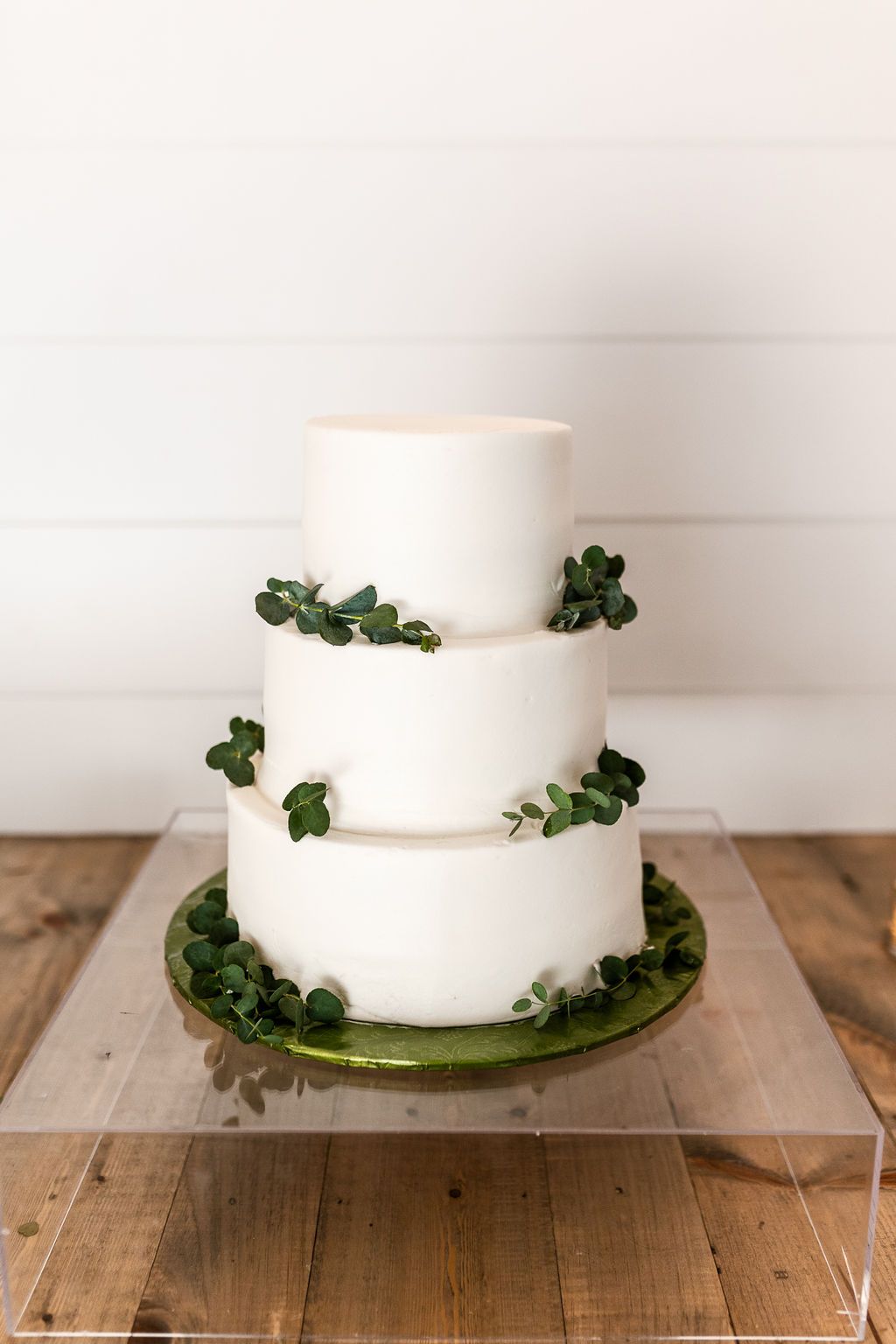 Three-tier white wedding cake decorated with fresh eucalyptus leaves on acrylic stand and wood table.