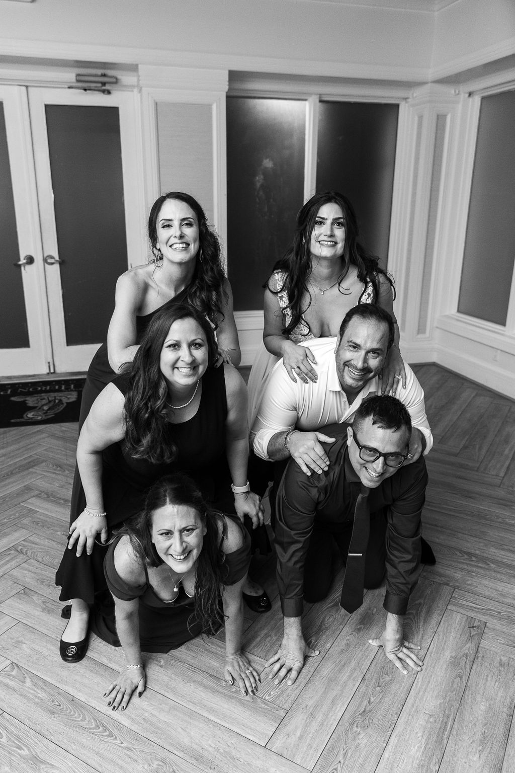 Black and white photo of five smiling wedding guests posing in a playful pyramid on the dance floor.