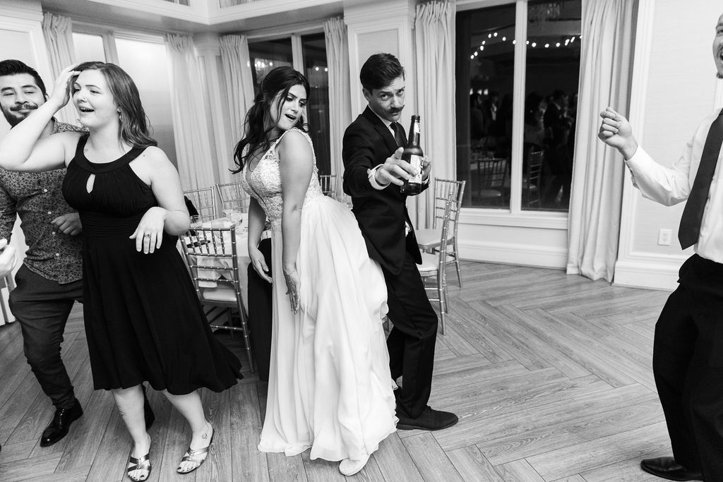 Black and white photo of wedding guests dancing joyfully at a reception, with bride in white gown.