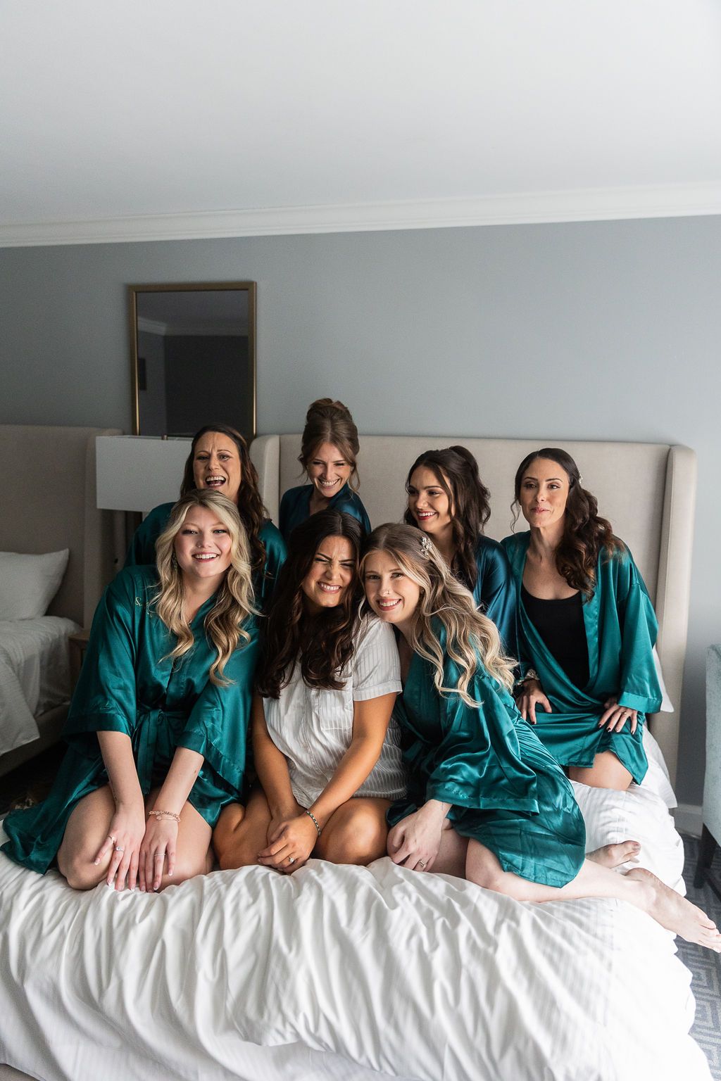 Bride and six bridesmaids in teal satin robes laughing and posing together on a white hotel bed.