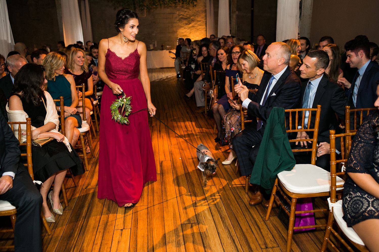 Bridesmaid in long burgundy dress walks down wooden aisle at elegant indoor wedding ceremony.