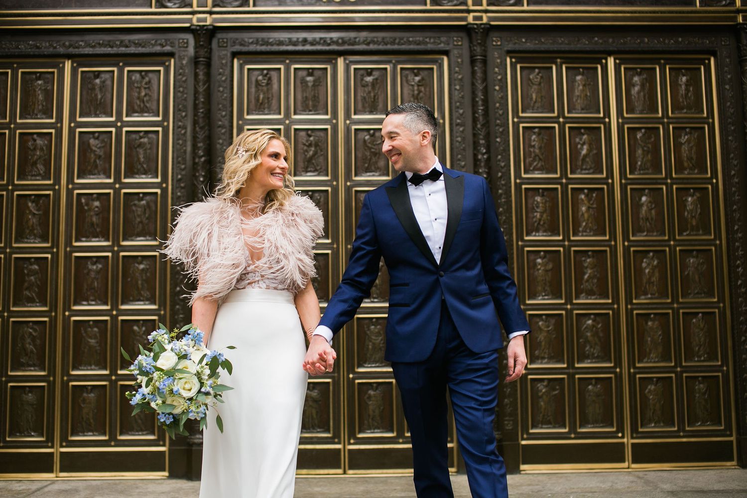 Bride and groom holding hands and smiling in front of ornate gold doors on their wedding day.