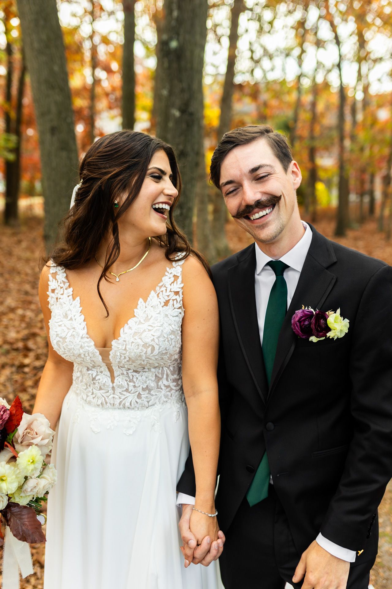 Laughing bride and groom with mustache share a joyful moment holding hands in autumn forest setting.