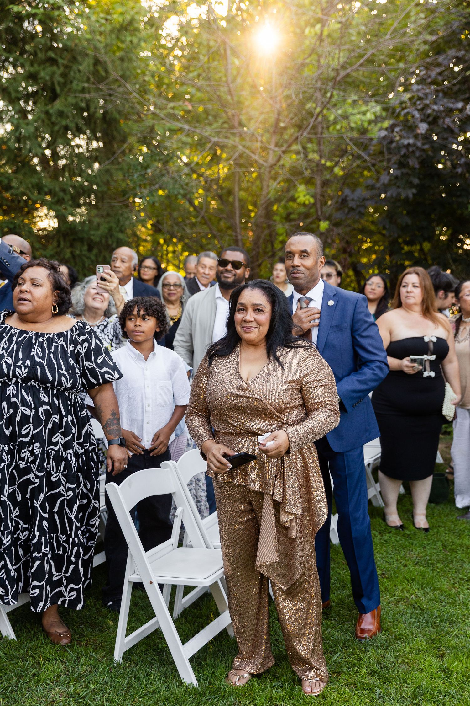 Wedding guests in formal attire stand on a lawn watching an outdoor ceremony at golden hour.