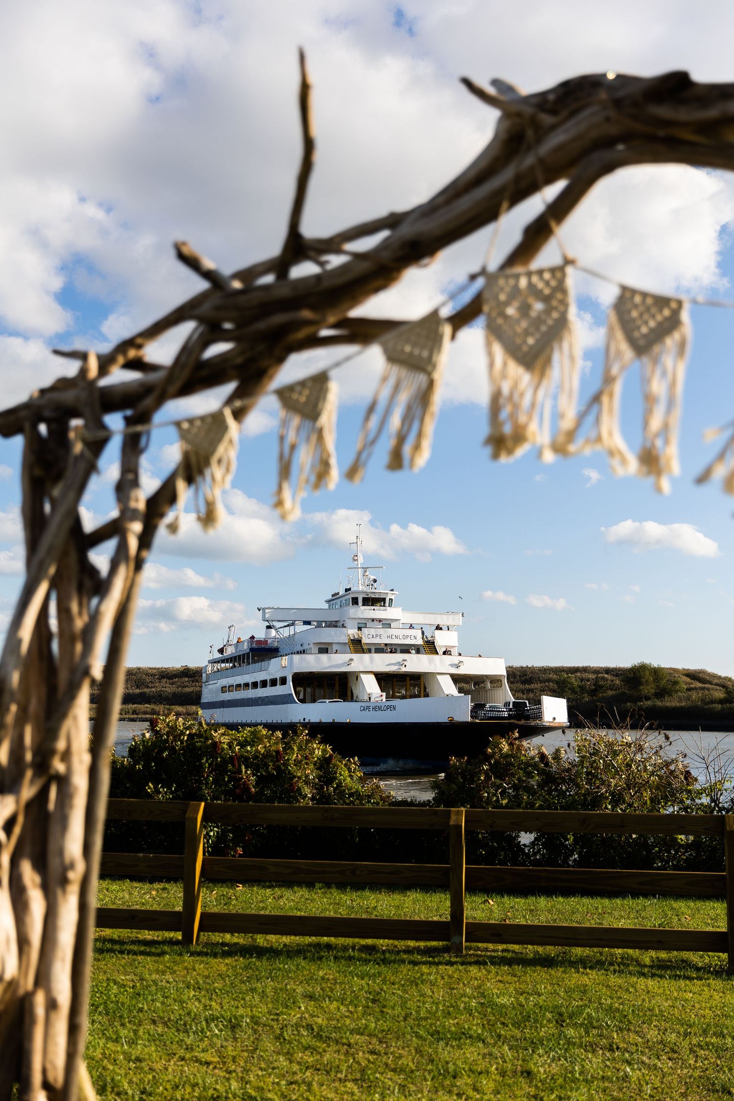 A driftwood arch with macrame bunting frames a white ferry boat on the water at a waterfront wedding venue.