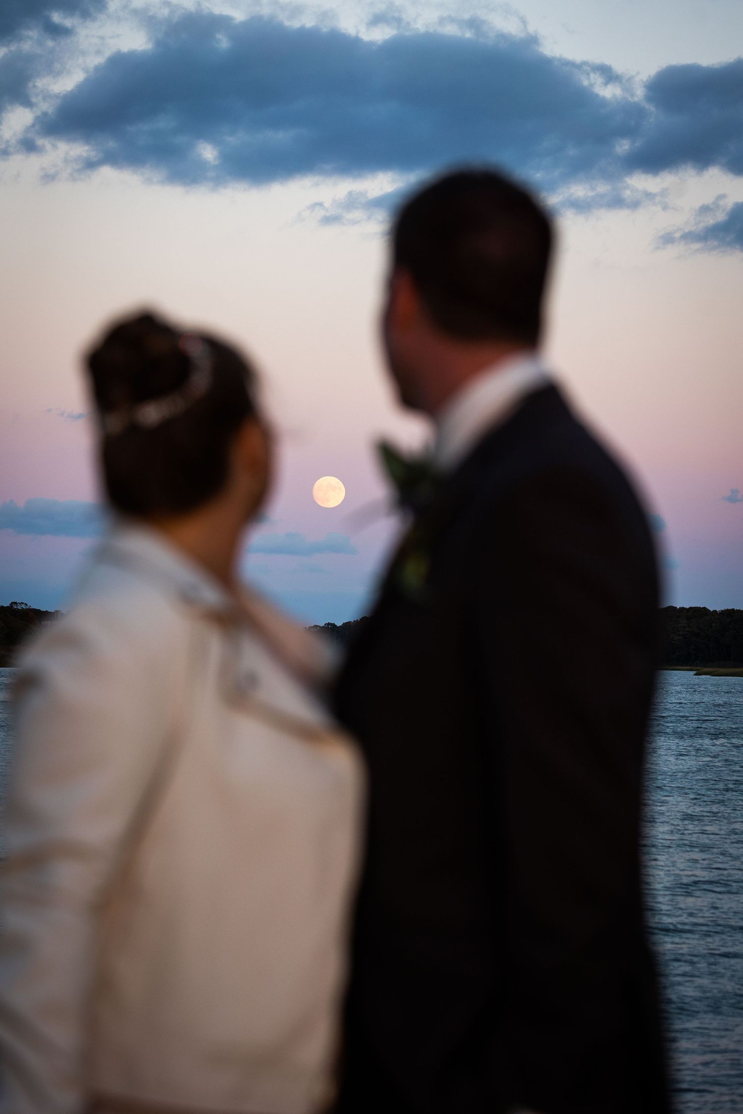 Bride and groom gazing at full moon rising over water at dusk after wedding ceremony.