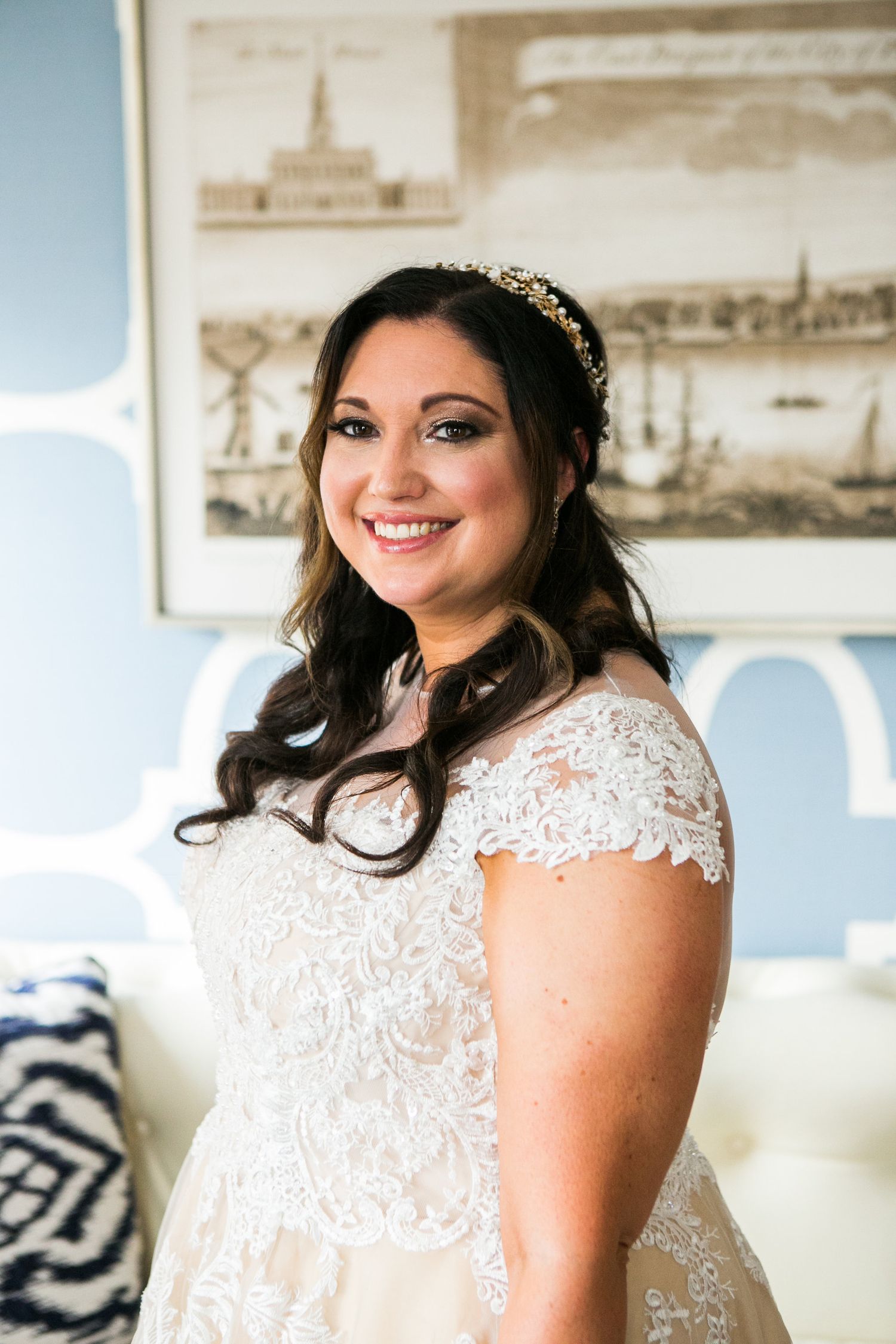 Radiant bride in a lace cap-sleeve wedding gown wearing a gold headpiece smiles warmly before her wedding.
