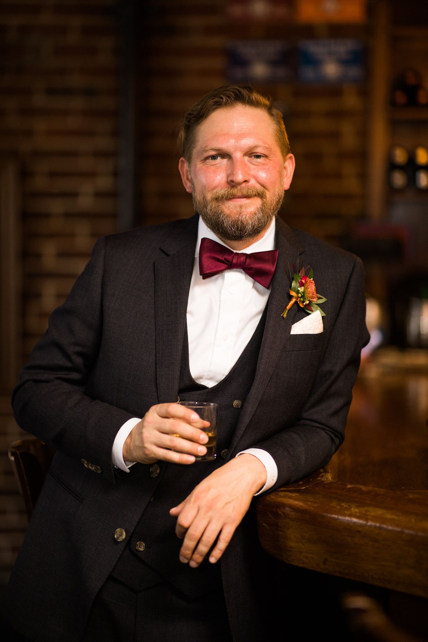 Handsome groom in dark suit with burgundy bow tie holding drink at bar.