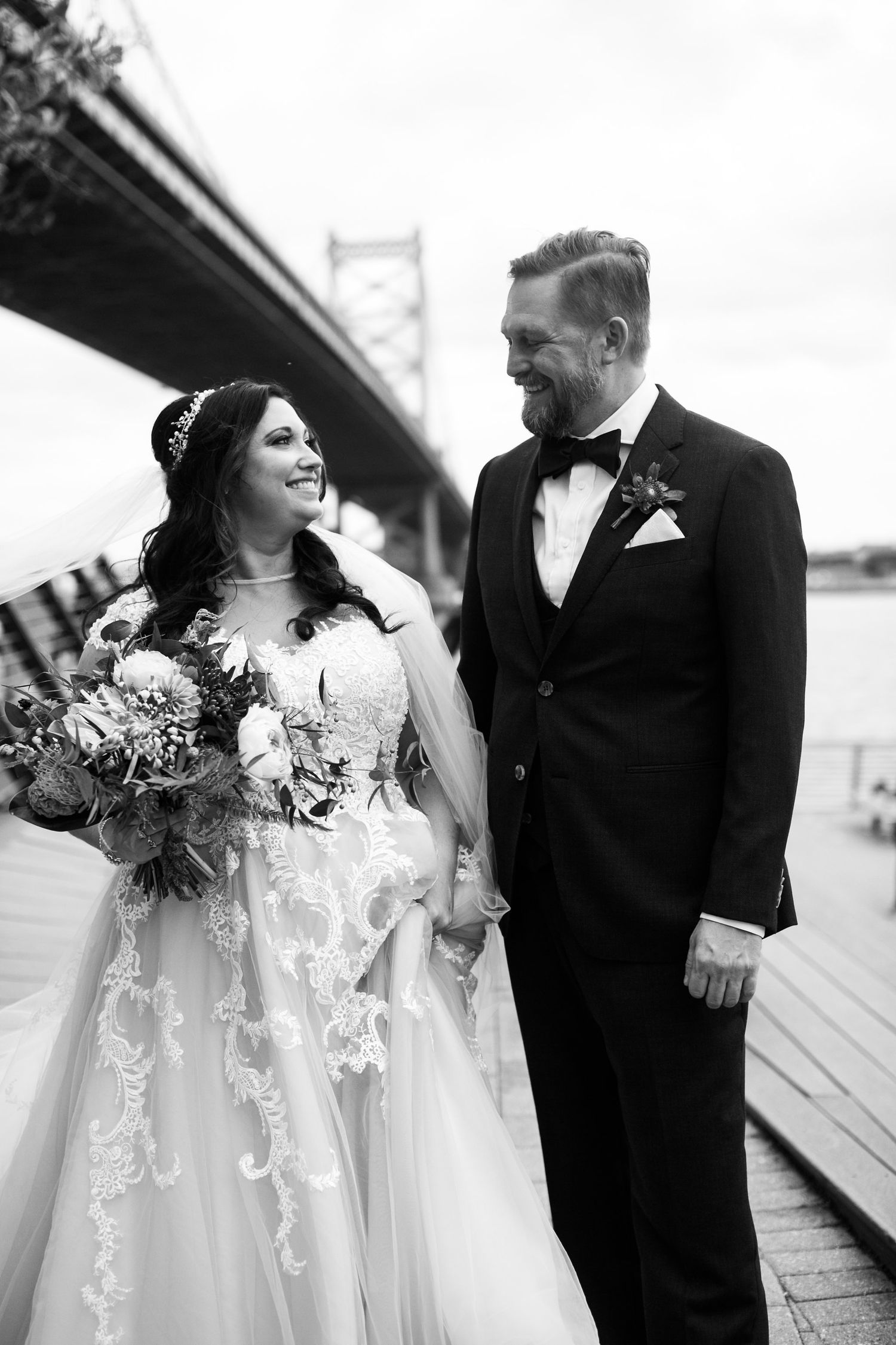 Black and white photo of bride and groom holding hands and smiling by riverside bridge.