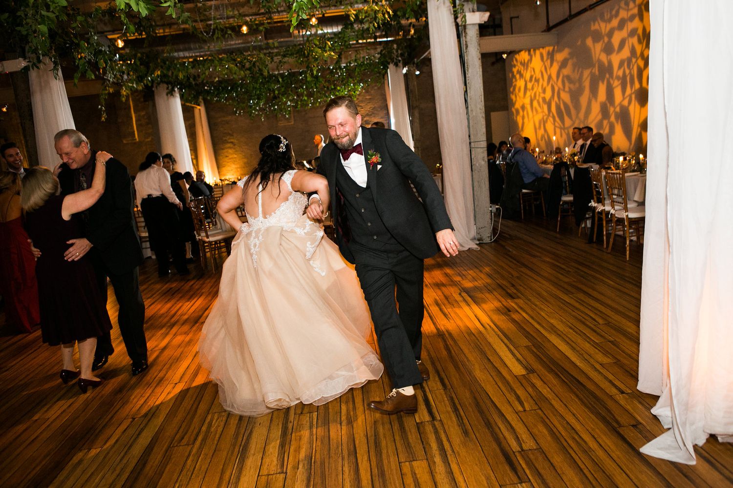 Bride in blush gown and groom in tuxedo dance on wooden floor at wedding reception.