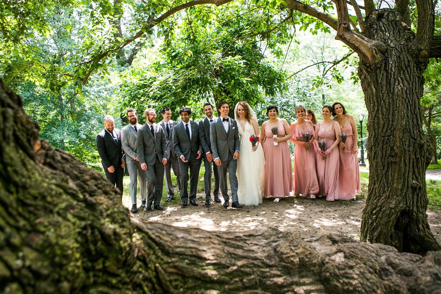 Wedding party in gray suits and pink bridesmaid dresses posing together under lush green trees.