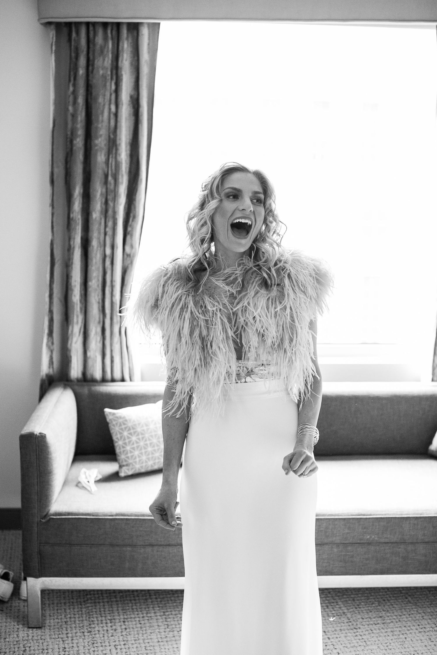 Black and white photo of excited bride in feather wrap laughing while getting ready.