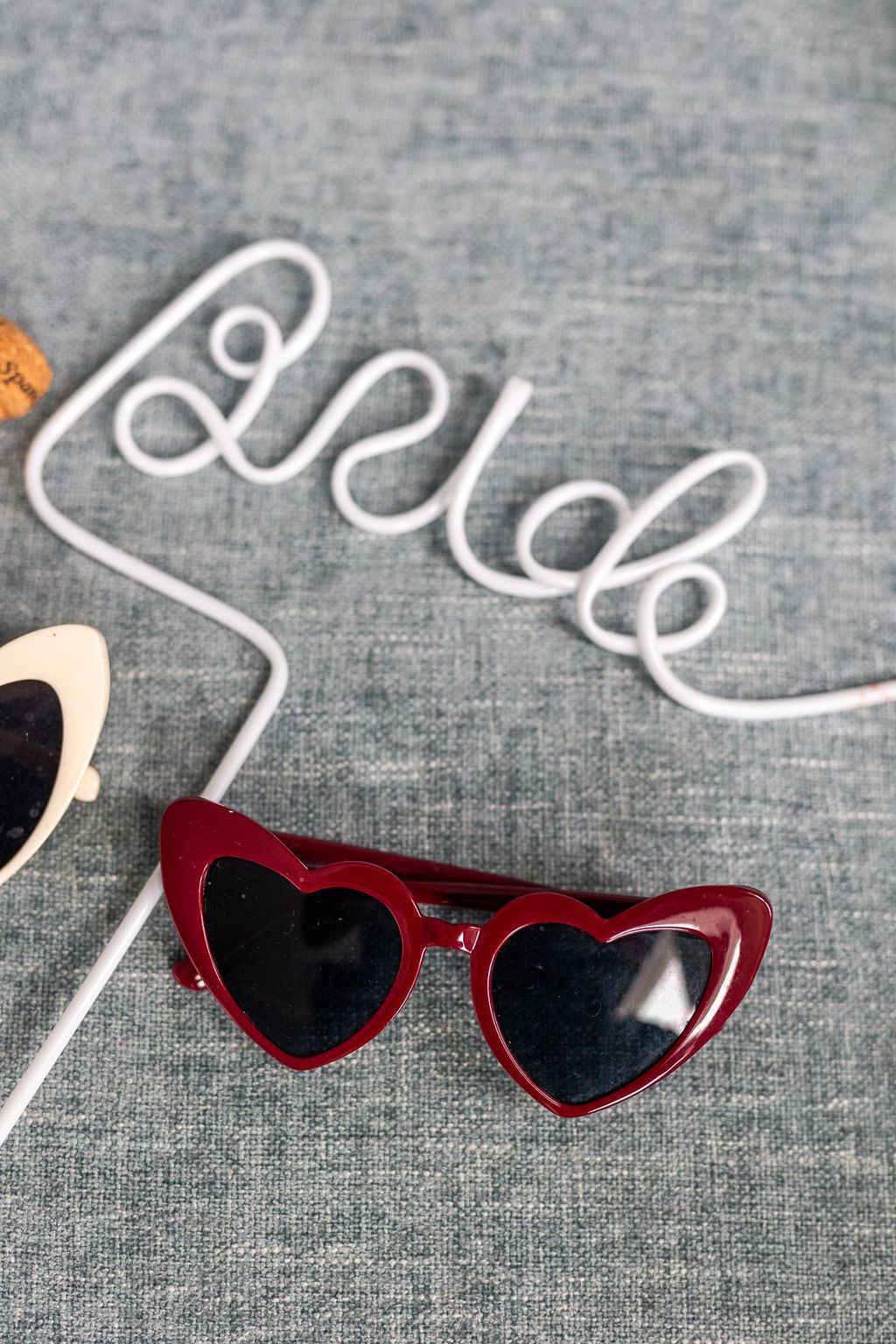 Red heart-shaped sunglasses beside a white wire Bride sign and white heart sunglasses on gray fabric.