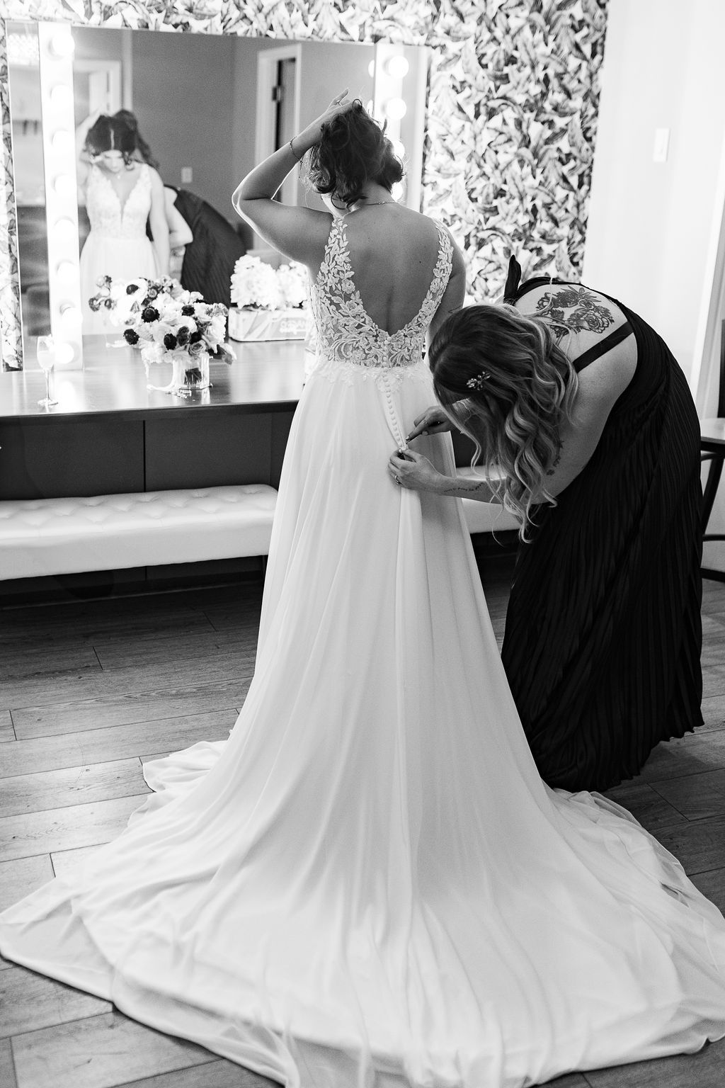 Black and white photo of bridesmaid fastening the back of a brides lace wedding gown in a bridal suite.