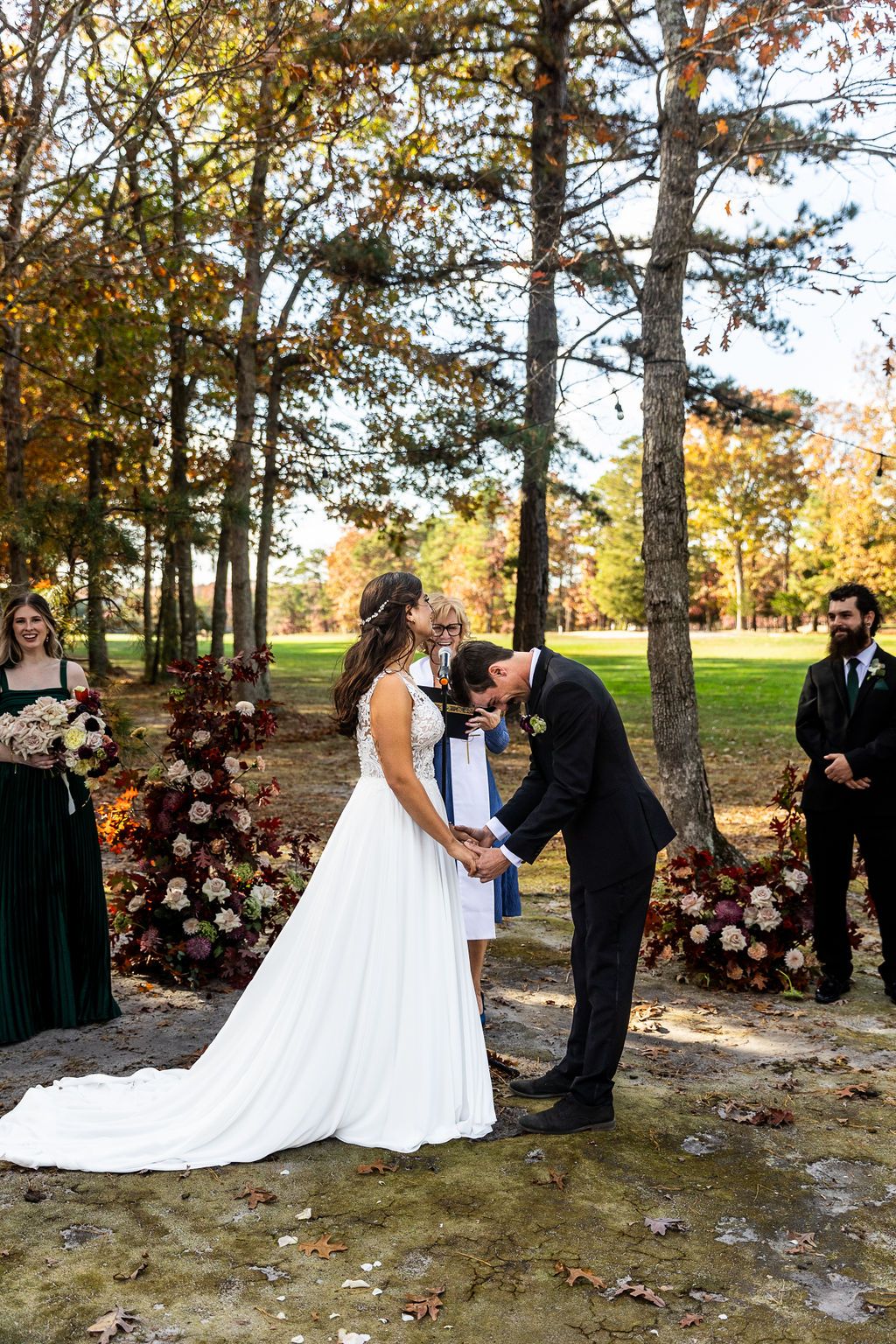 Groom bows his head during outdoor fall wedding ceremony while bride stands beside him holding hands.