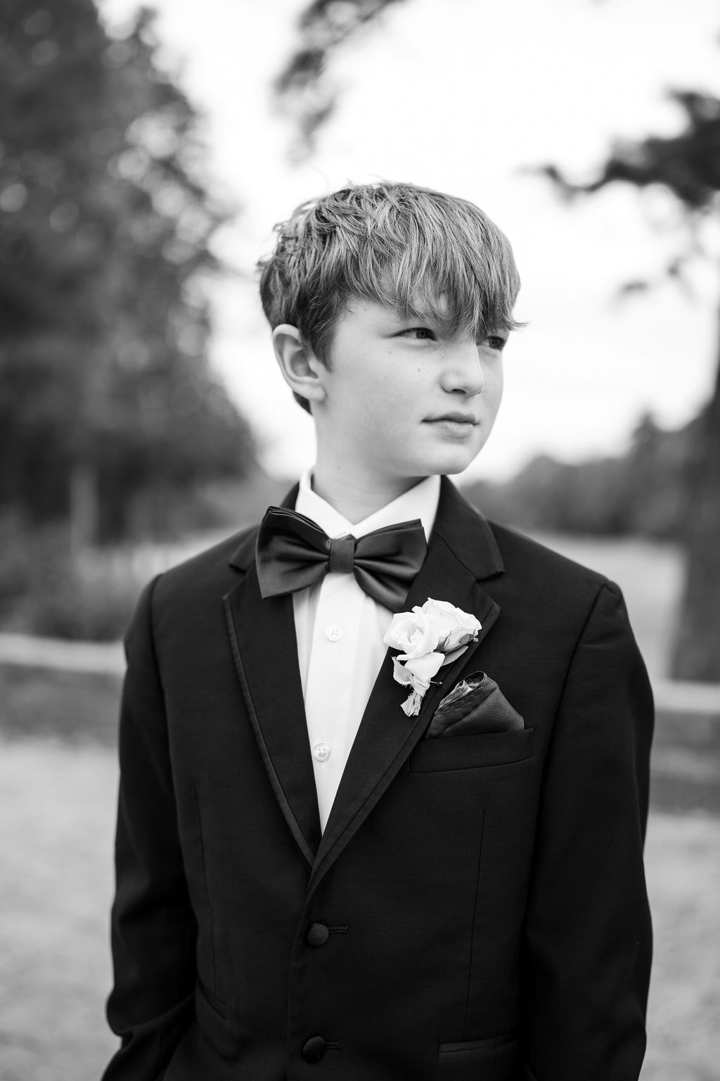 Young ring bearer in black tuxedo with bow tie and boutonniere poses outdoors in black and white.