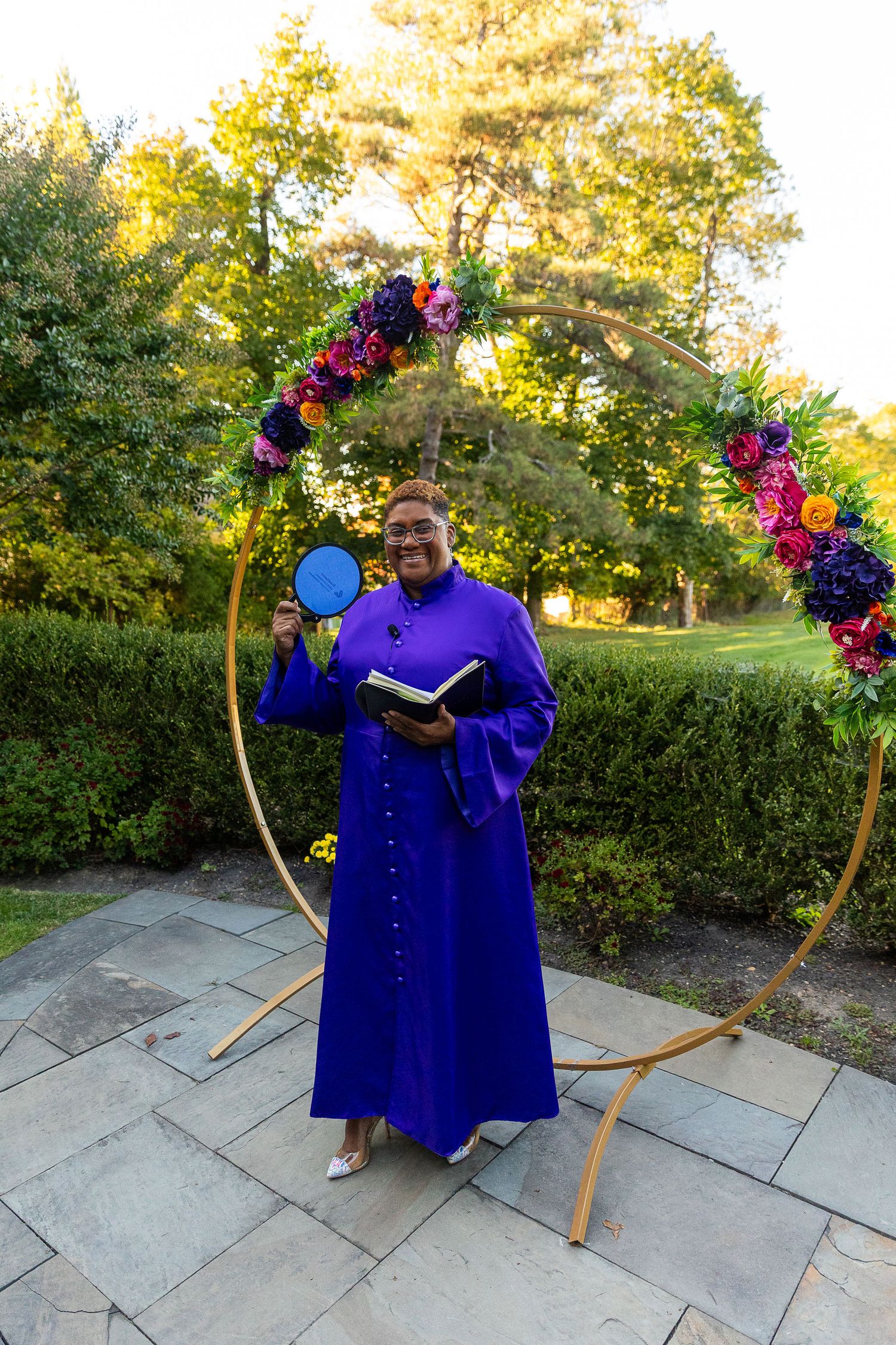 Officiant in purple robe smiling before floral arch at outdoor wedding ceremony.