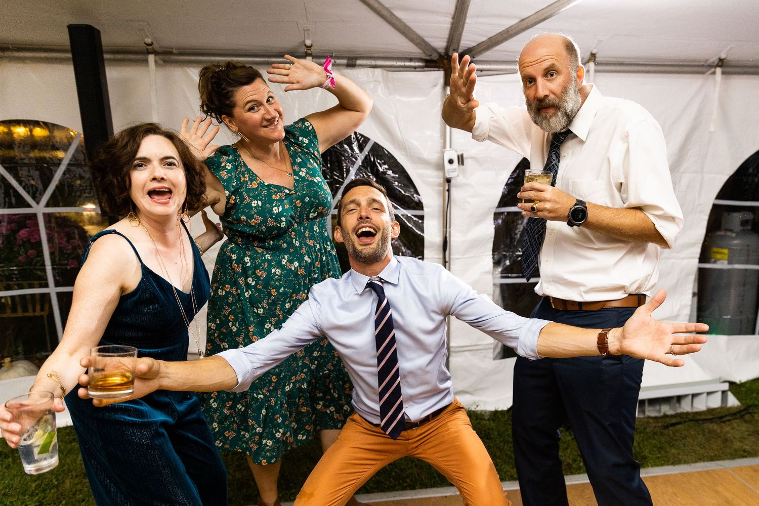 Four wedding guests dance and laugh together energetically under a tent at a reception.