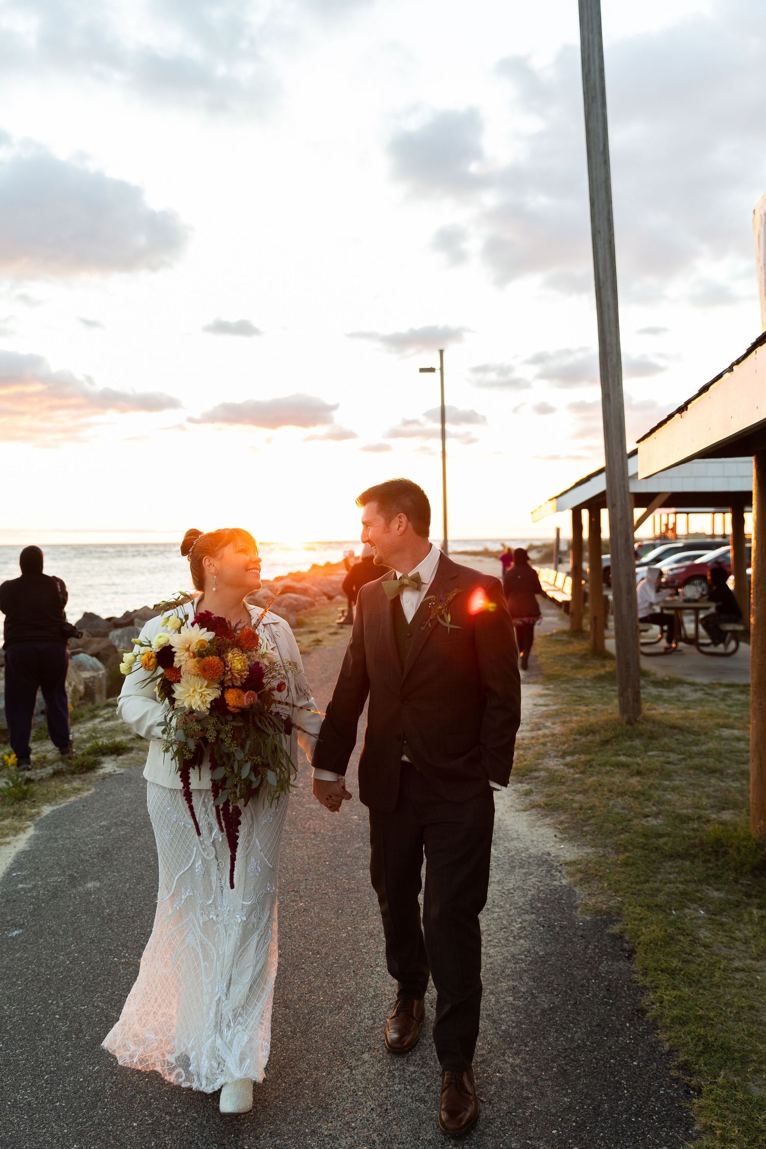 Bride and groom walking hand in hand along coastal path at sunset with colorful bouquet.