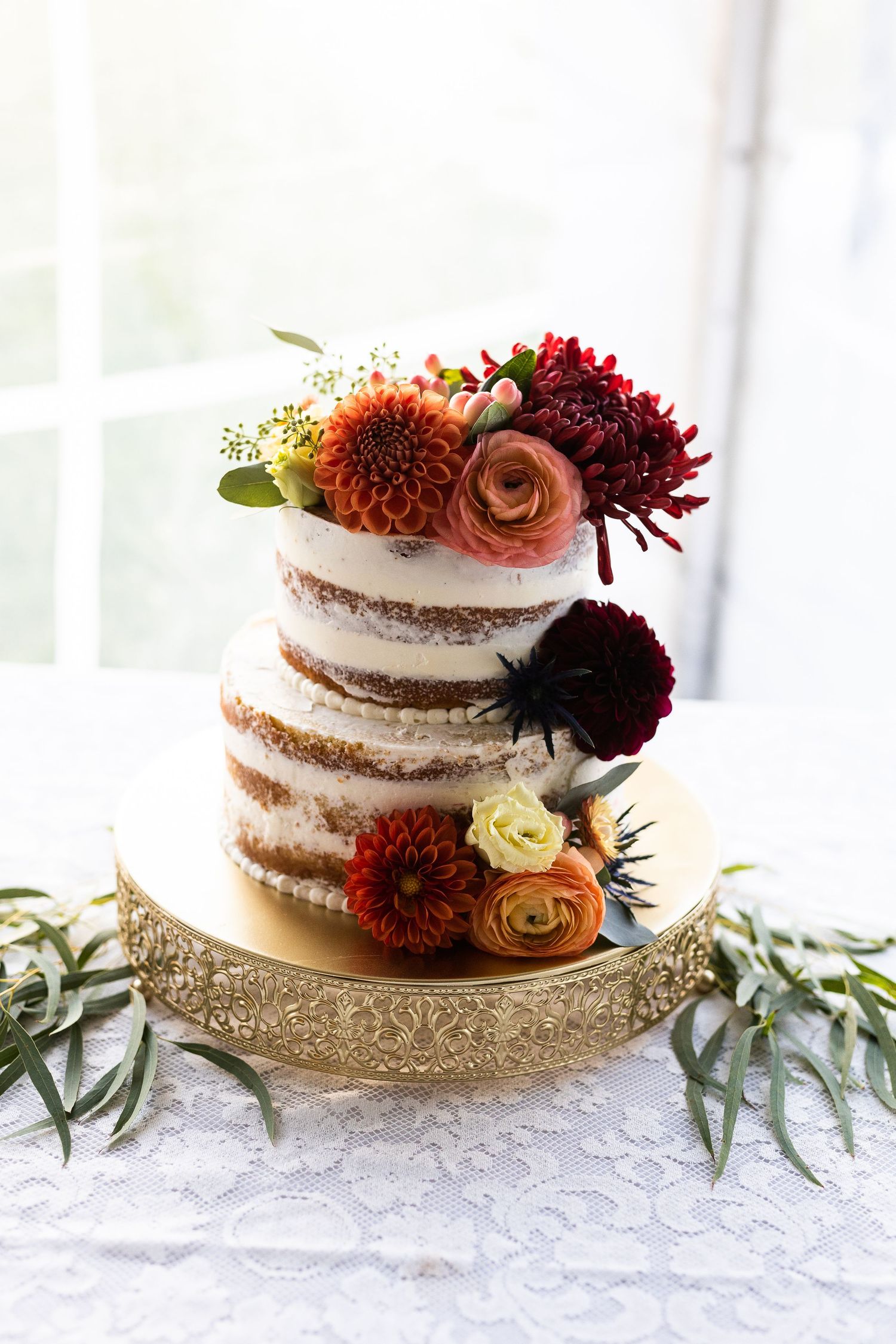 Elegant two-tier naked wedding cake adorned with fall dahlias and ranunculus on gold stand.