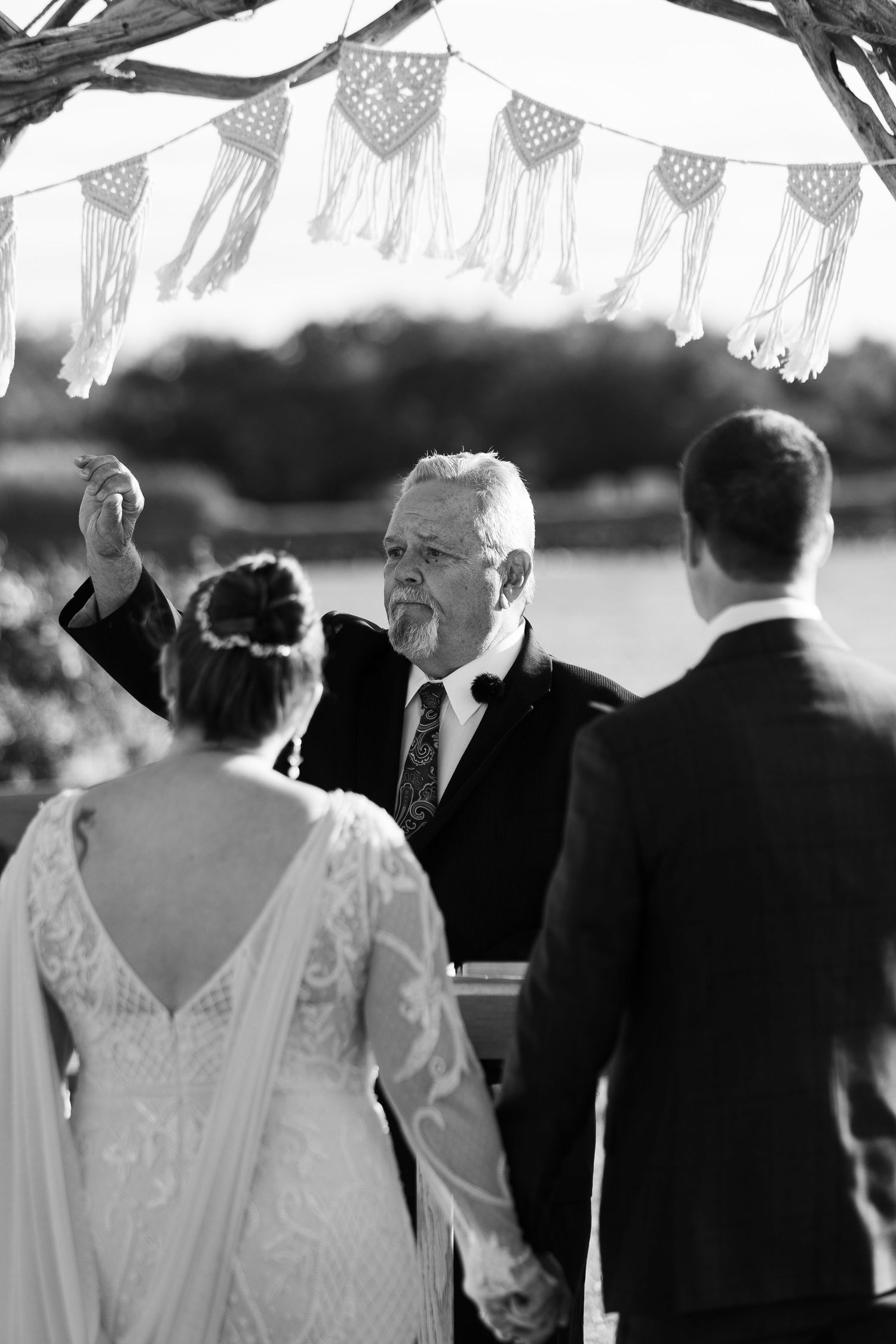 Black and white photo of officiant raising hand during outdoor wedding ceremony under macrame arch.