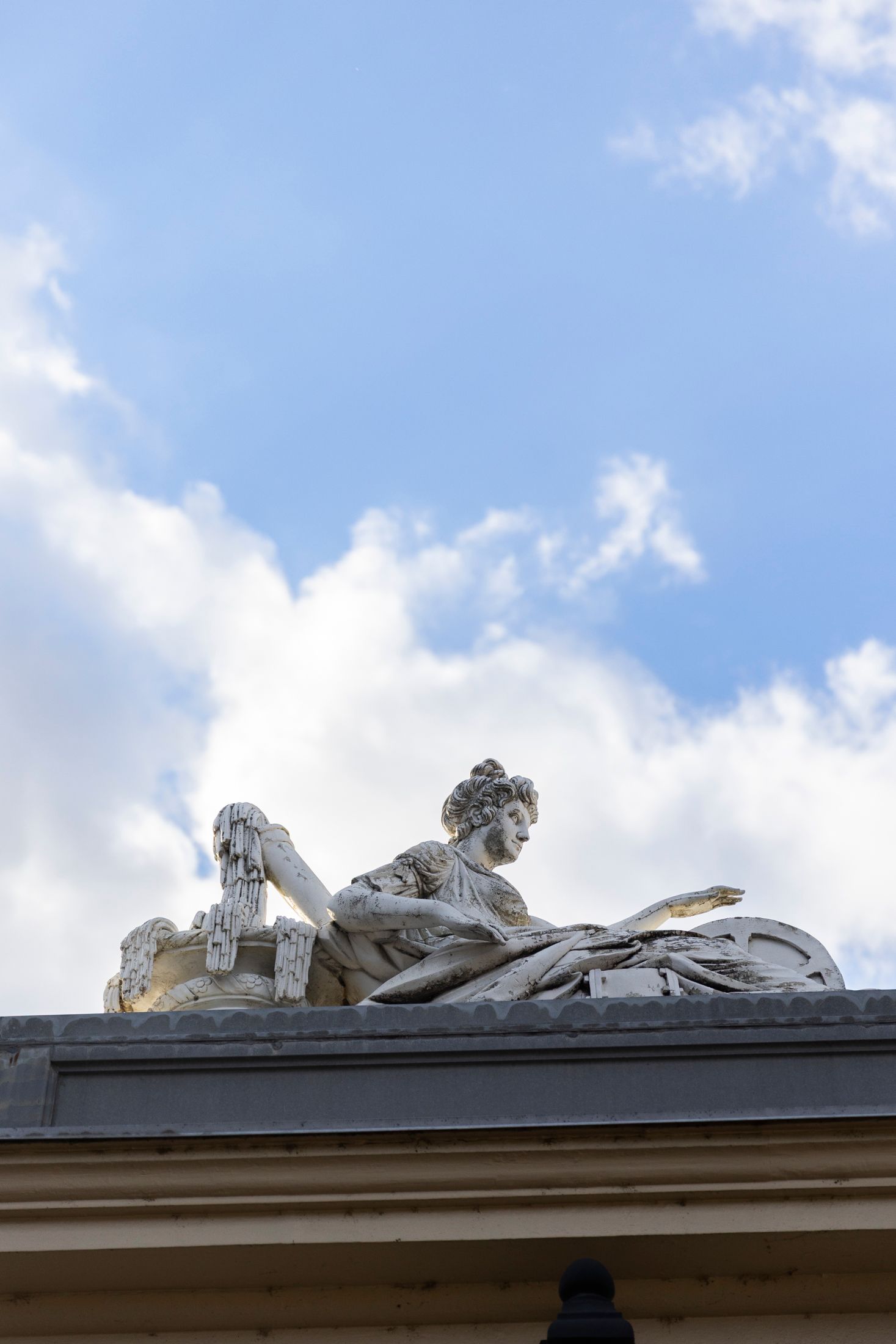 Classical white stone reclining figure sculpture atop a building against a partly cloudy sky.