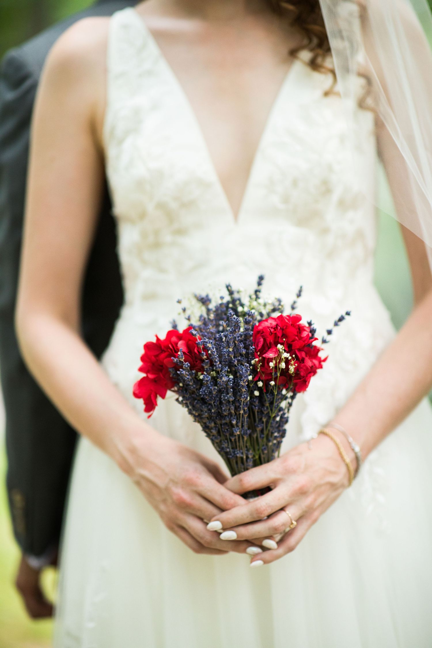 Bride in a white lace V-neck gown holds a small bouquet of red blooms and purple lavender.