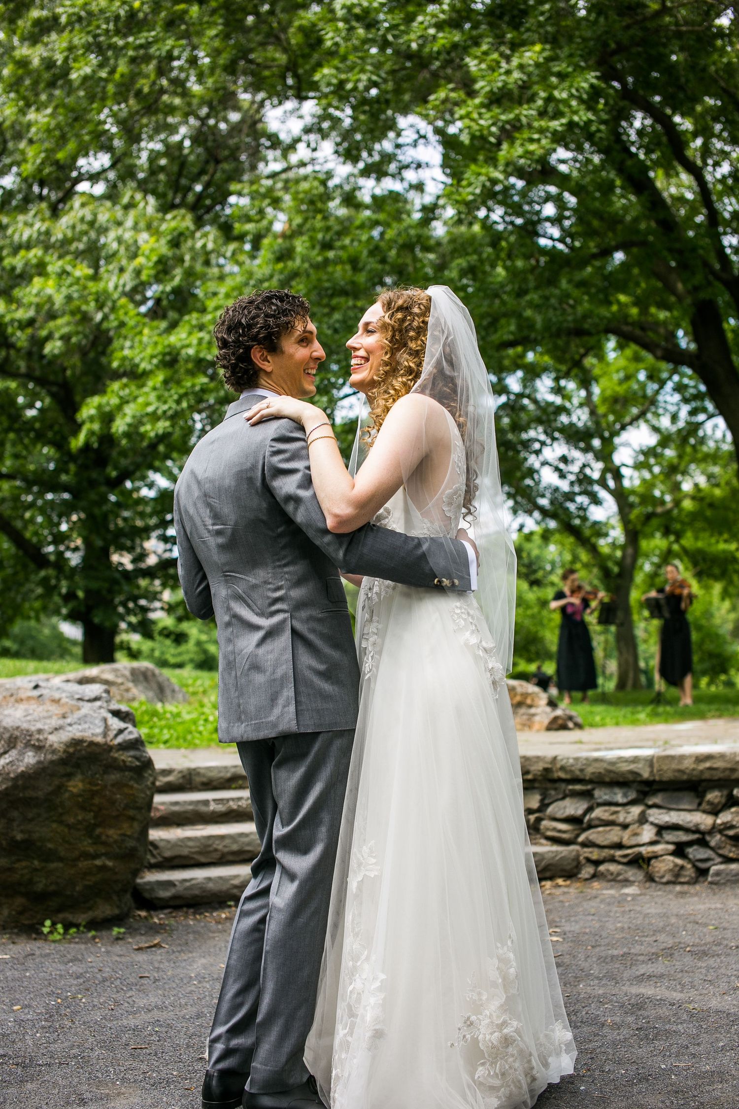 Bride in white veil and groom in gray suit share a romantic embrace and smile at outdoor wedding.