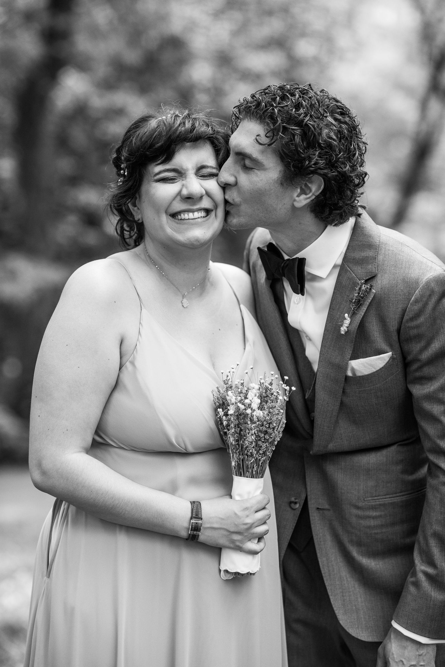 Black and white portrait of a groom kissing a smiling woman on the cheek at a wedding celebration.