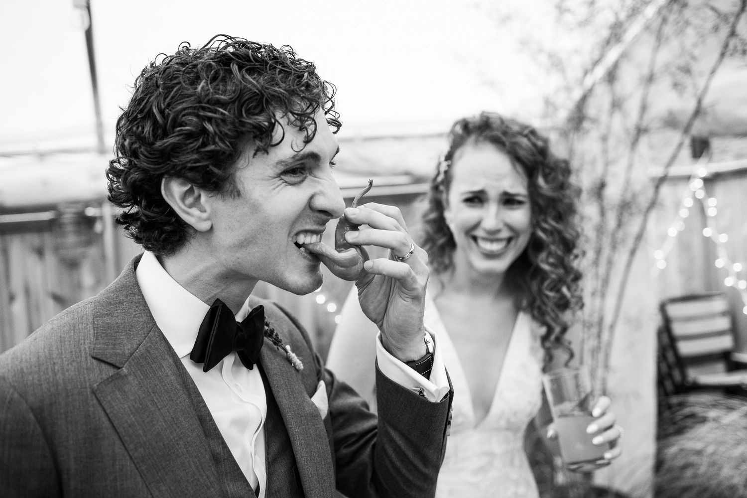 Black and white photo of groom eating wedding cake while bride laughs joyfully beside him.