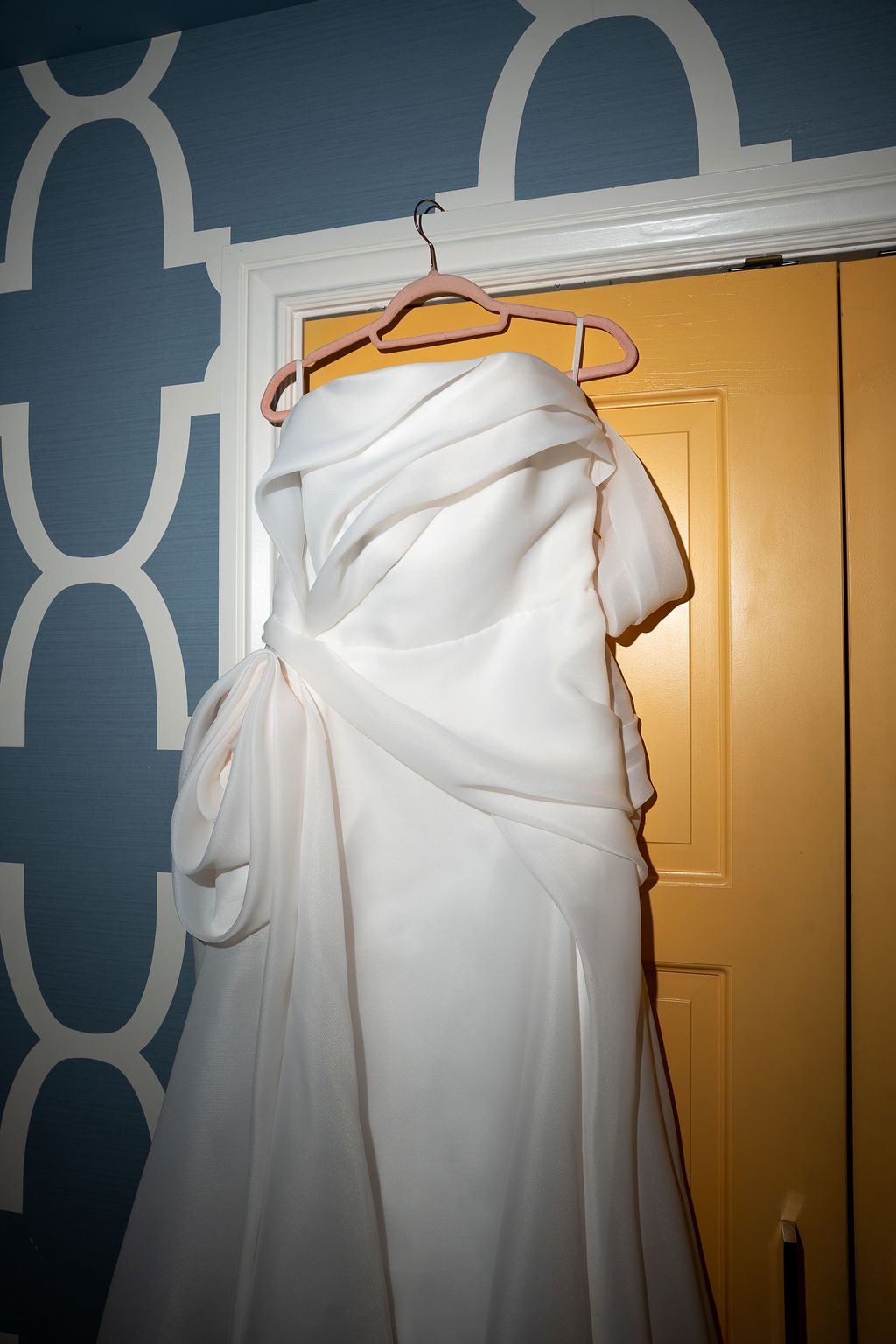 Elegant white strapless wedding gown with draped bow detail hanging on a hanger against a blue patterned wall.