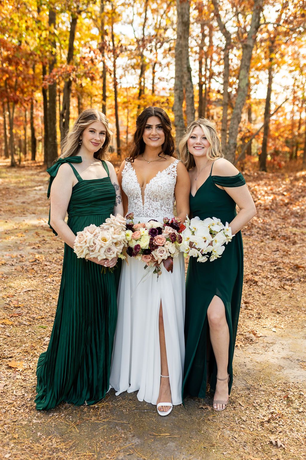 Bride in white lace gown flanked by two bridesmaids in emerald green dresses holding white bouquets in autumn woods.