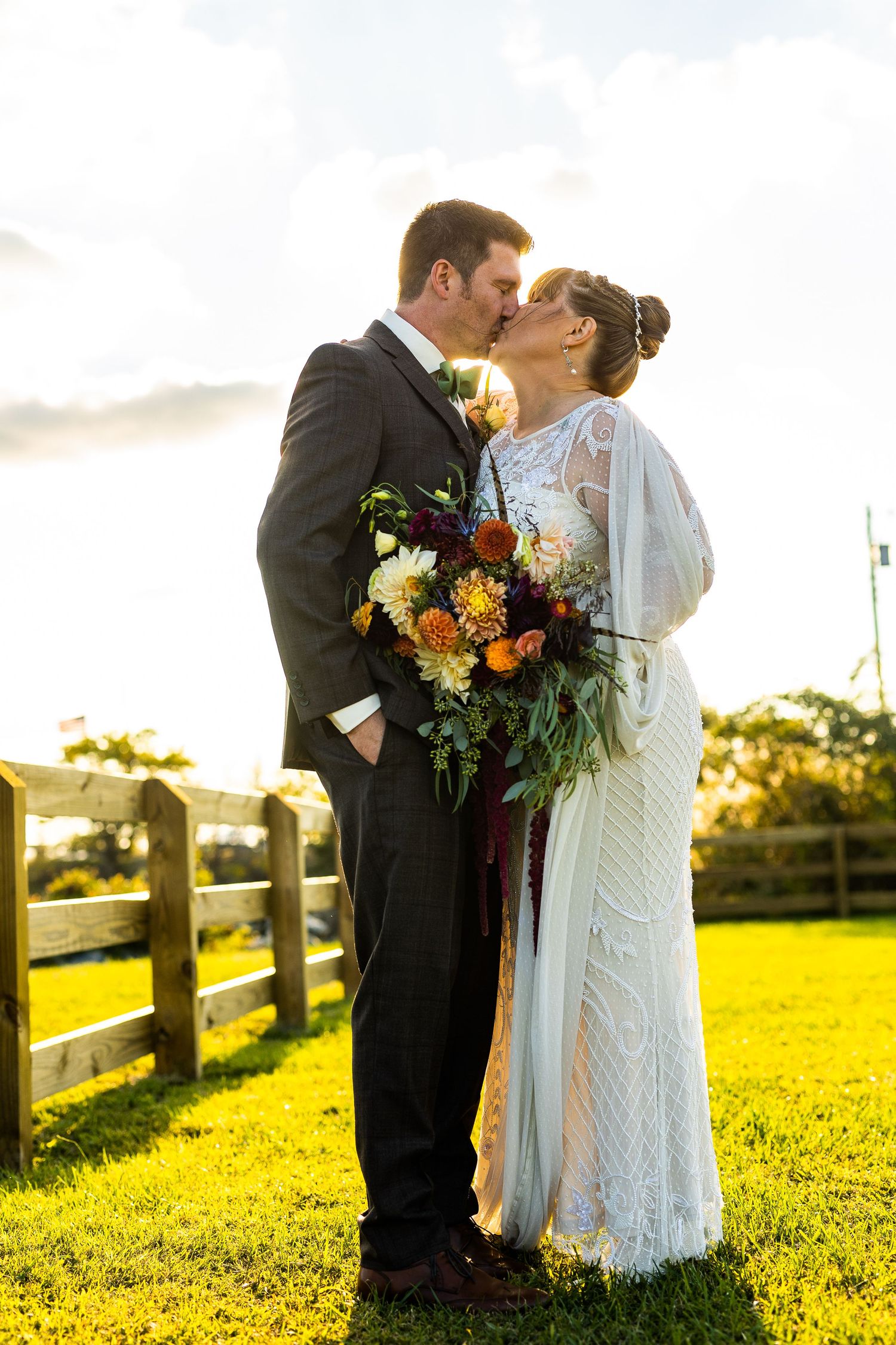 Bride and groom sharing a kiss at sunset, bride holding a vibrant autumn bouquet by a wooden fence.